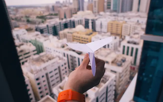 Hand holding a white paper airplane above a blurred city view