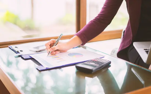Hand writing on financial charts beside a calculator on a glass office desk